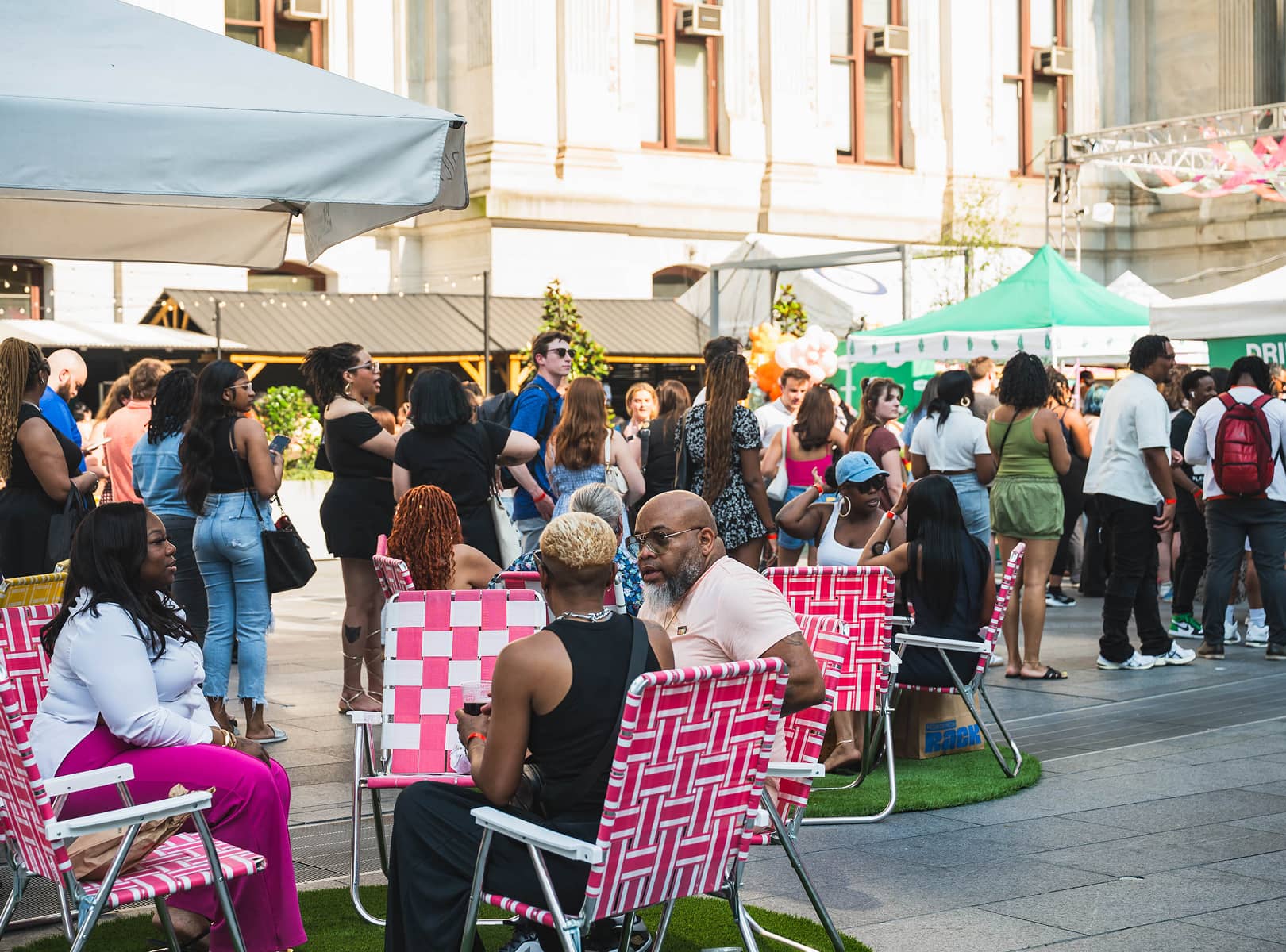 People gather at an outdoor event in a city square. The scene is lively, with groups chatting, colorful tents, and pink chairs, creating a festive atmosphere.