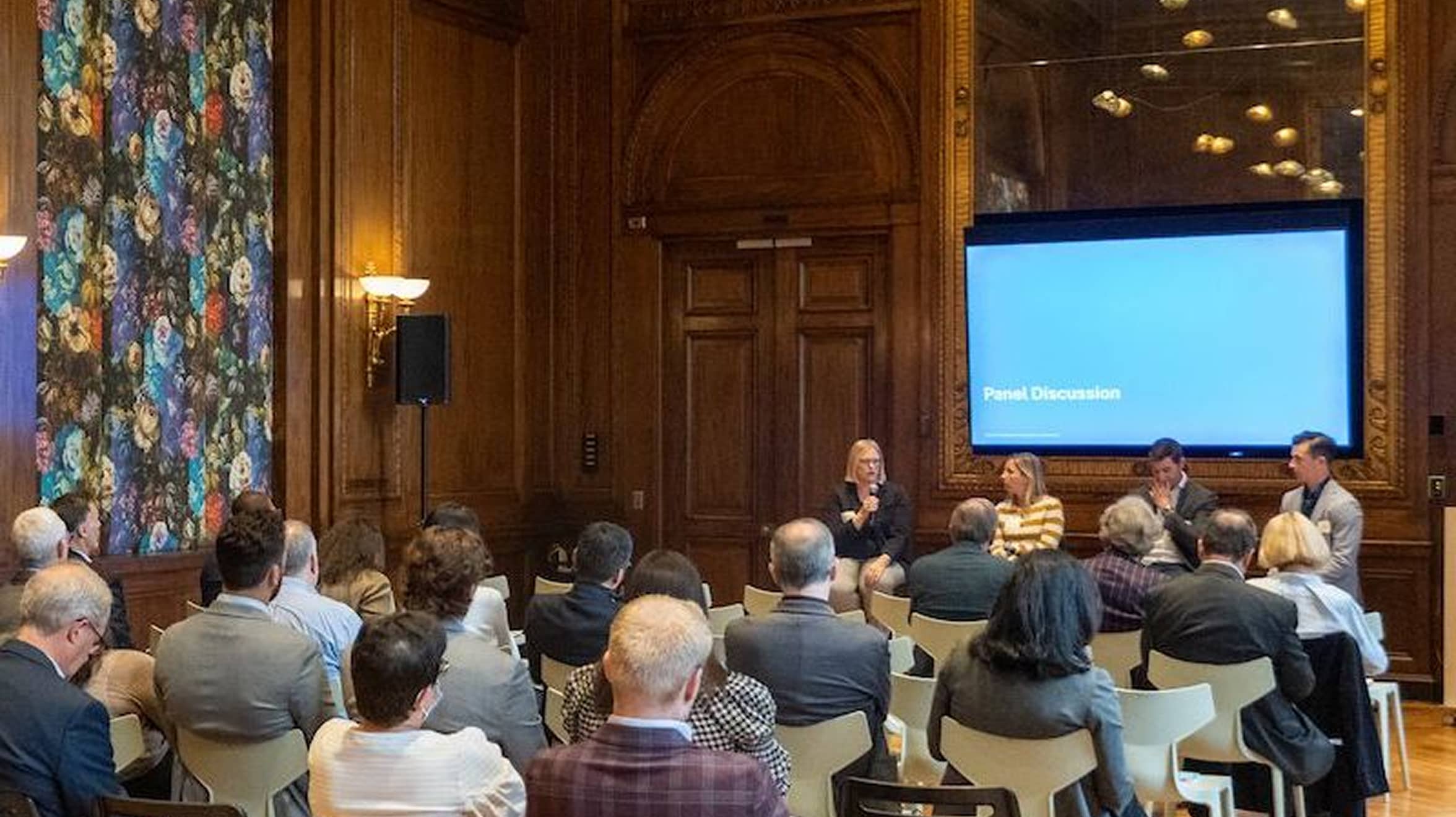 Audience seated in an elegant wood-paneled room with floral artwork, facing a panel discussion on stage. A speaker addresses the group, conveying a professional tone.