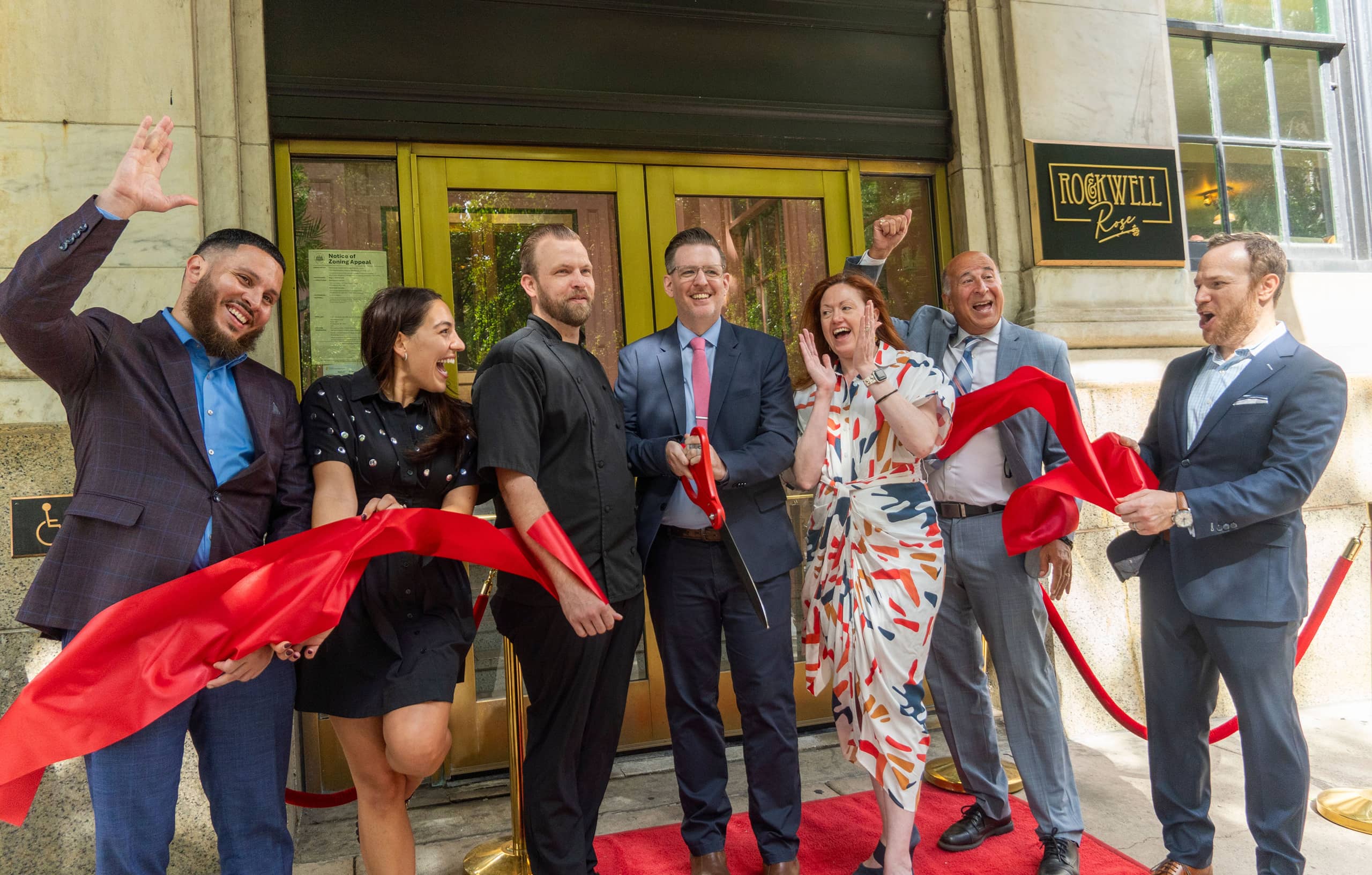 A group of seven joyful people stands in front of a building entrance, cutting a red ribbon. They are celebrating, smiling broadly, and wearing formal attire.