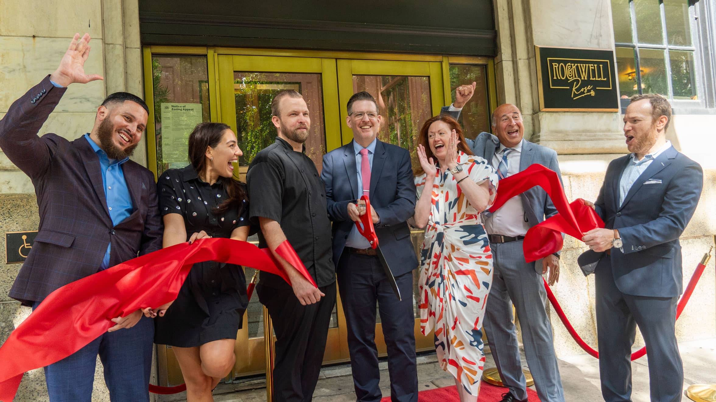 A group of seven joyful people stands in front of a building entrance, cutting a red ribbon. They are celebrating, smiling broadly, and wearing formal attire.