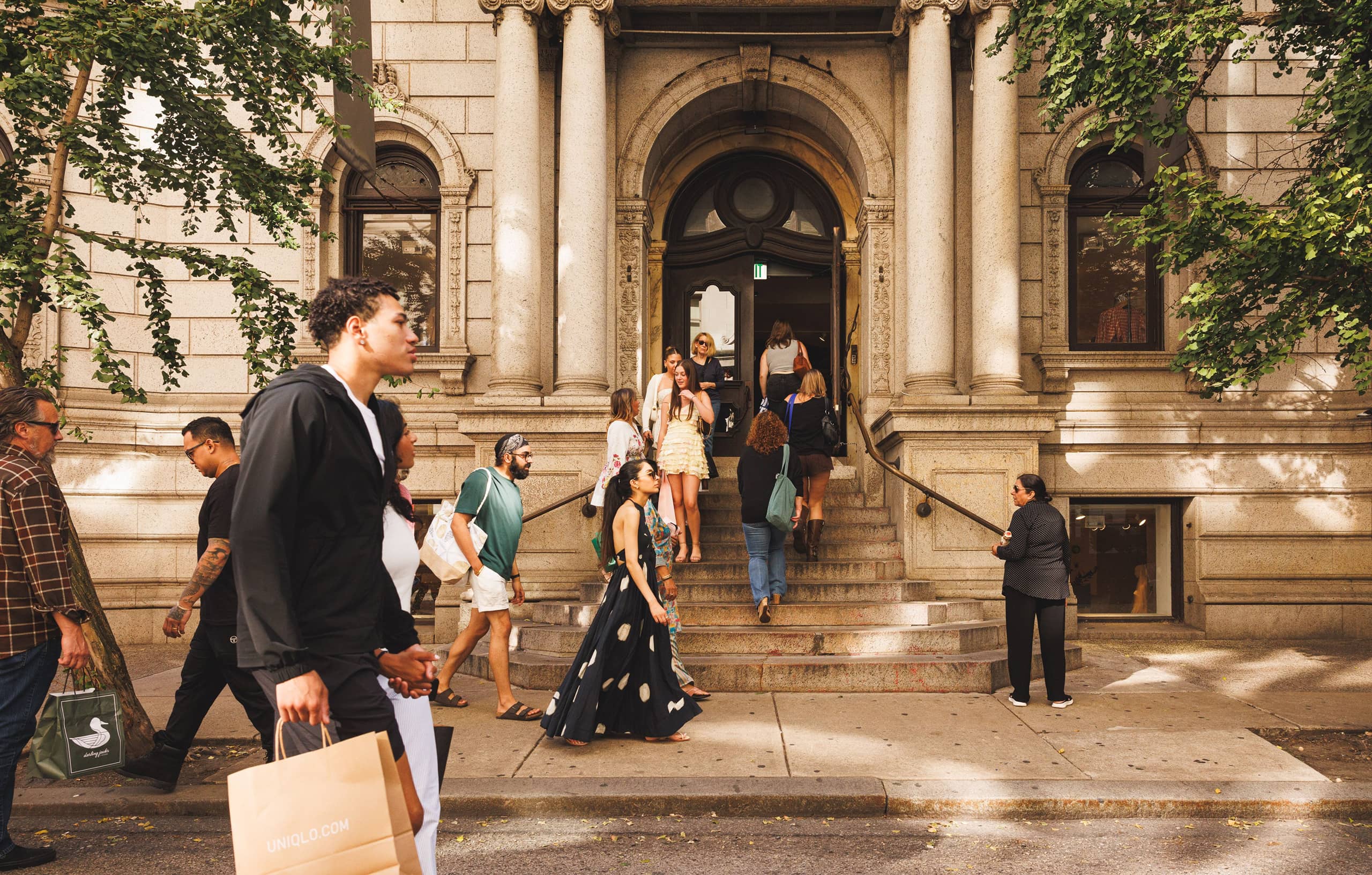 Pedestrians walk past a grand building entrance where people are ascending the steps. Some carry shopping bags. The scene is lively and sunny.