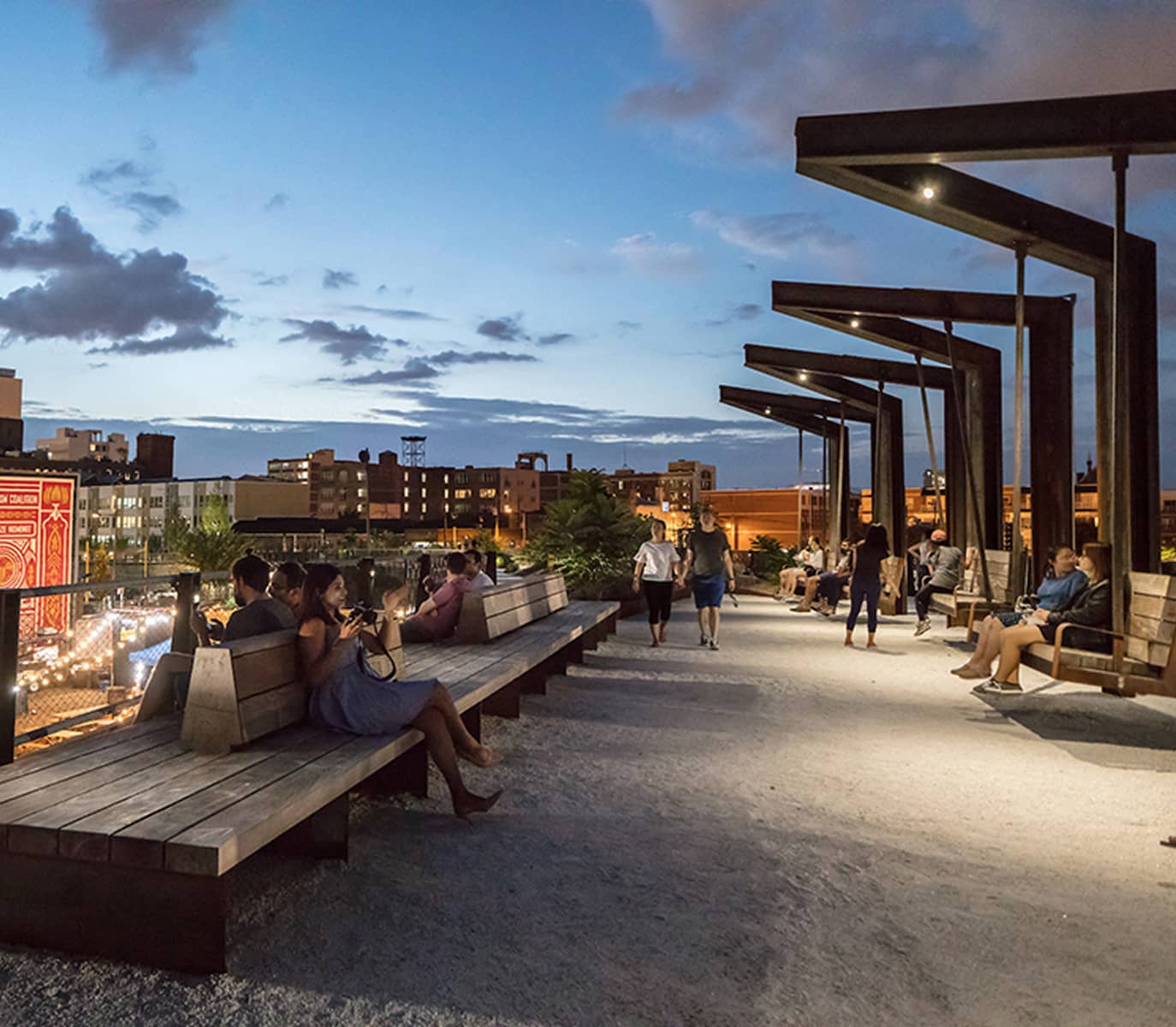 People relax on wooden benches under modern black pergolas at a park during twilight. The sky glows softly, creating a calm, urban ambiance.