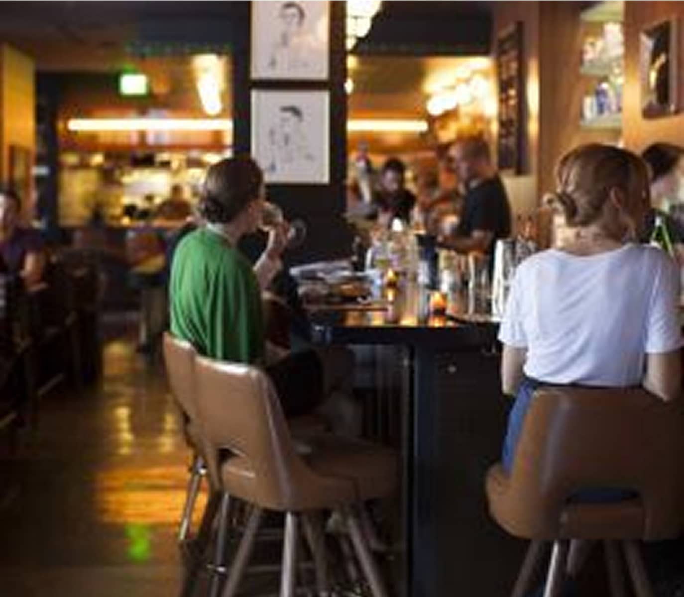 A cozy, dimly-lit café with people seated at a bar. A woman in a white shirt and another in green are shown from behind, sipping drinks. Warm, inviting ambiance.