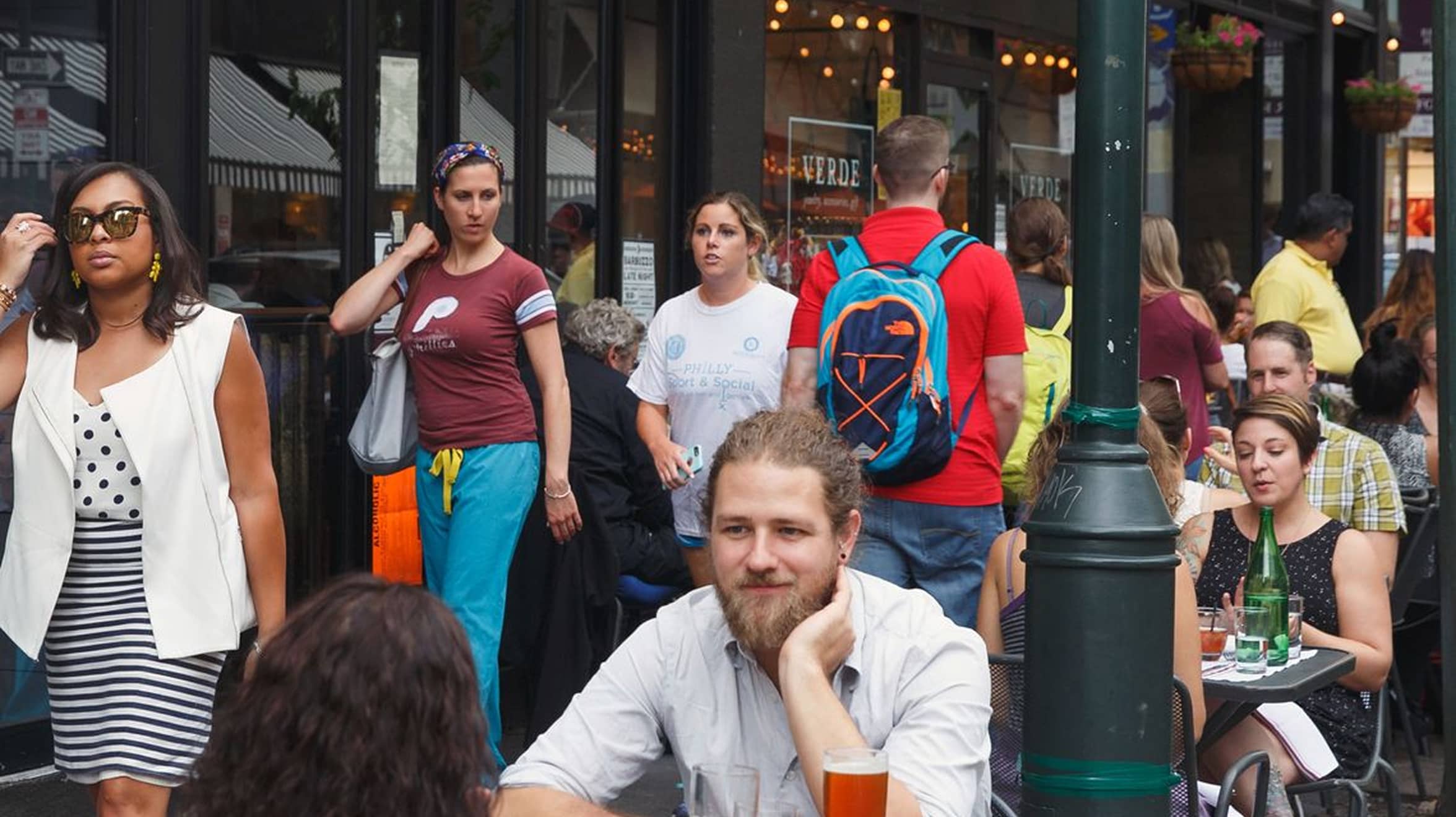 Outdoor cafe scene with a relaxed bearded man at a table with drinks, surrounded by bustling pedestrians and diners. Casual and lively atmosphere.
