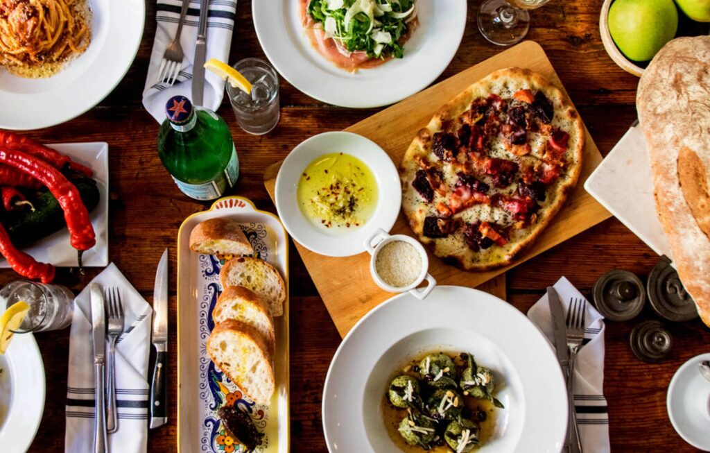 A wooden table set with various dishes, including pizza, sliced bread, salad, and pasta. Vibrant and cozy dining setting with a welcoming and appetizing atmosphere.
