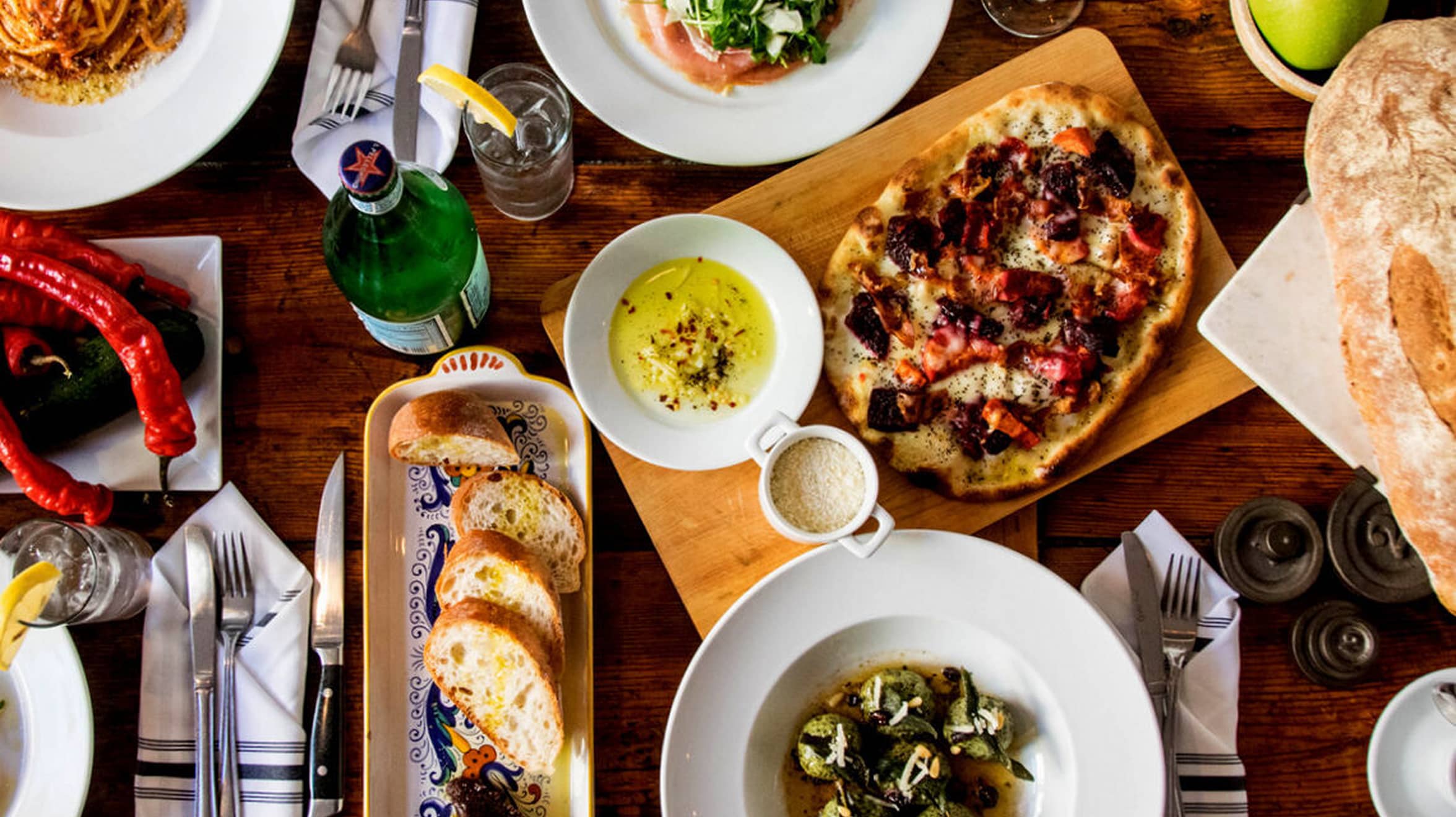 A wooden table set with various dishes, including pizza, sliced bread, salad, and pasta. Vibrant and cozy dining setting with a welcoming and appetizing atmosphere.
