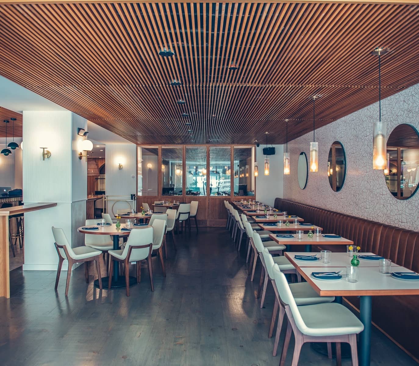 Modern restaurant interior with wooden ceiling and floor, featuring neatly arranged tables and white chairs. Warm lighting and mirrors create an inviting atmosphere.