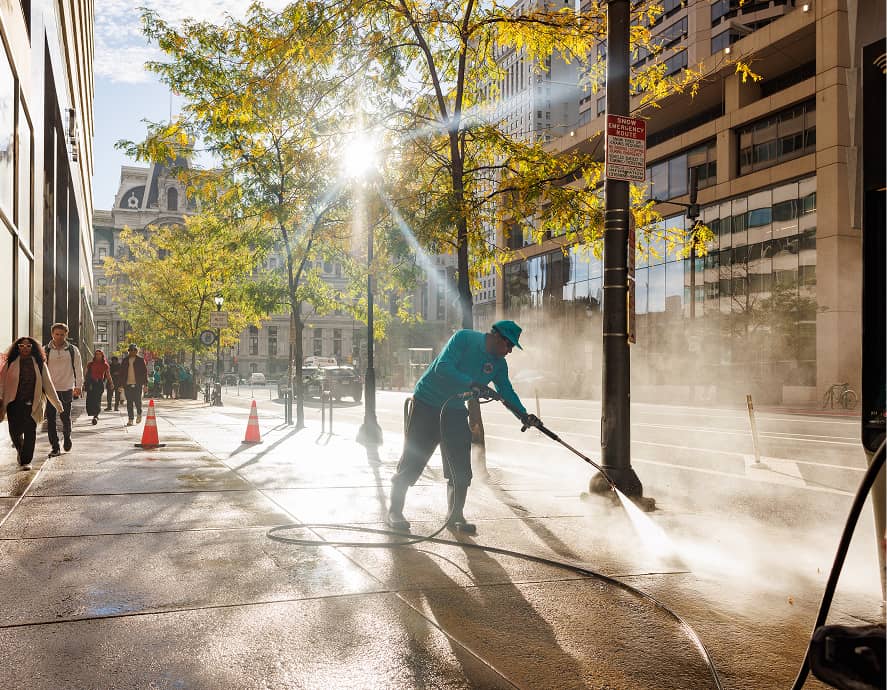 A CCD worker in blue uses a hose to power wash a city sidewalk. Sunlight filters through trees, casting shadows. People walk by, framed by tall buildings.