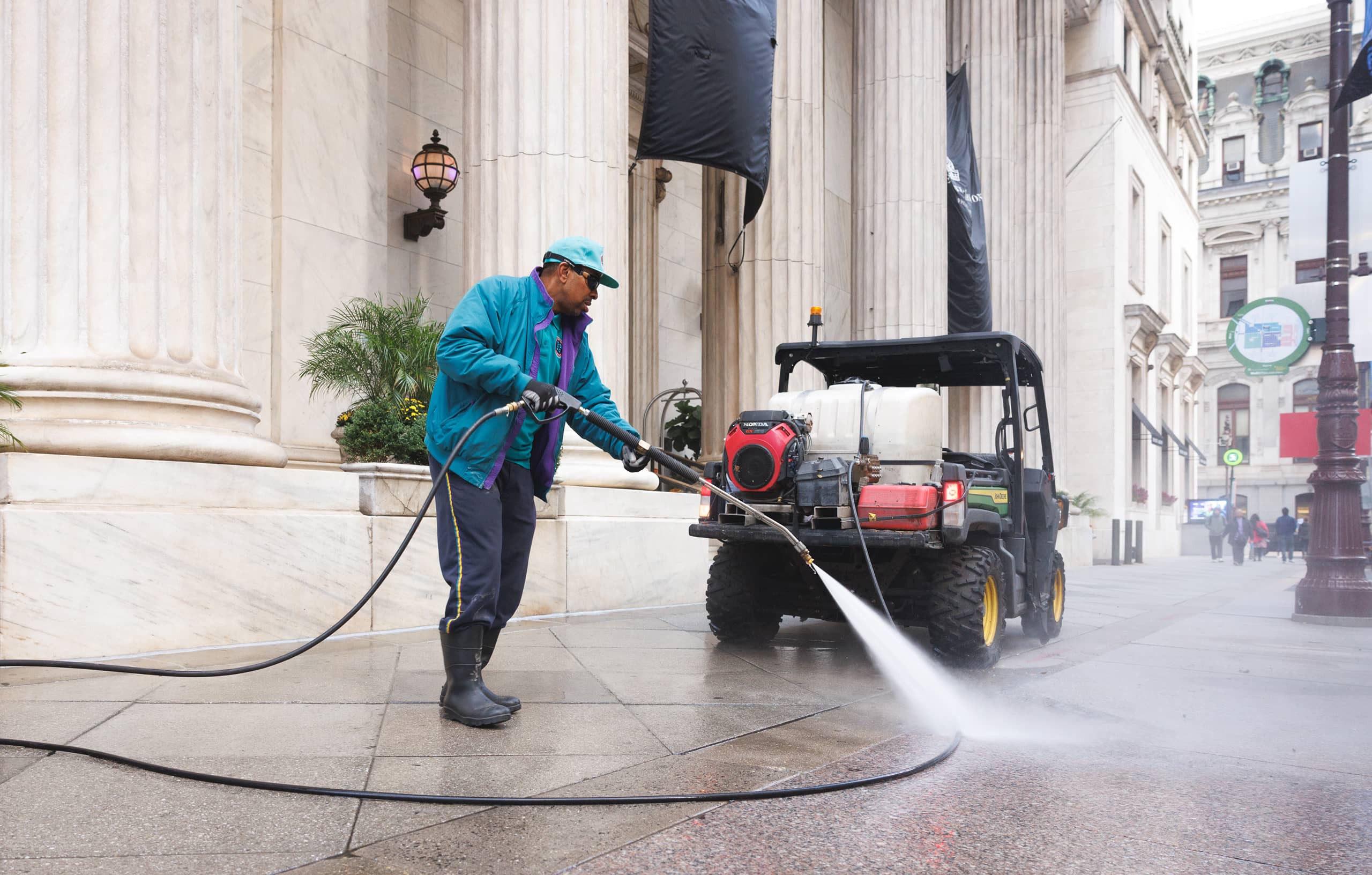 A person in teal and blue clothing power washes a sidewalk in front of a grand building with columns. A utility vehicle is parked nearby.