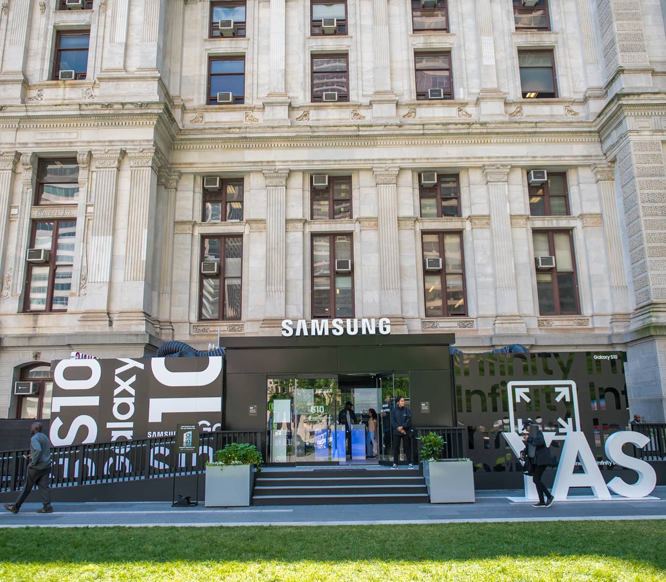 Facade of a grand building with Samsung signage and large Galaxy S10 displays. People enter a sleek, modern store, creating a bustling, tech-savvy atmosphere.