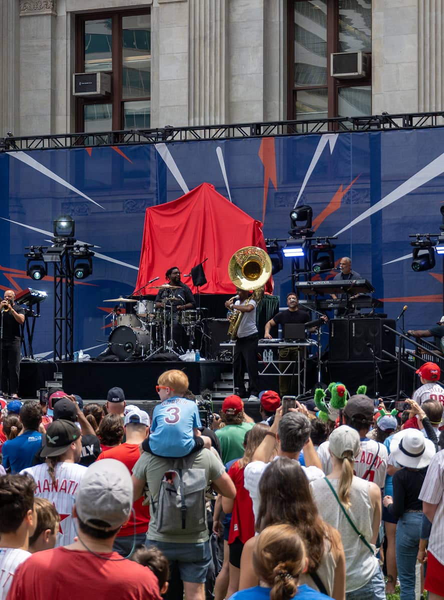 A vibrant outdoor concert scene with a diverse crowd watching musicians on stage. A tuba player stands out, conveying a lively, festive atmosphere.