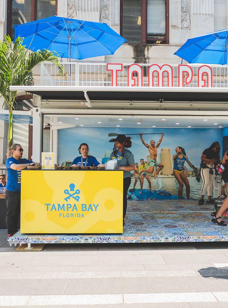 Outdoor Tampa Bay Florida kiosk with yellow signage and employees. Blue umbrellas and palm trees add a tropical vibe; backdrop shows people enjoying the beach.