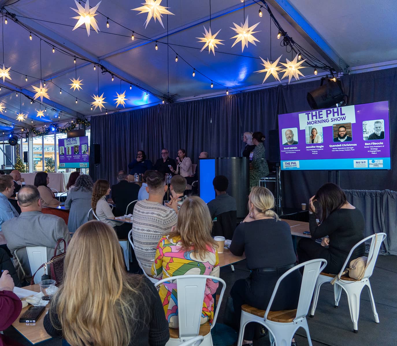 A busy panel discussion takes place under a tent with star-shaped lights and blue ambient lighting. Attendees sit at tables, focused on the speakers. A screen displays 