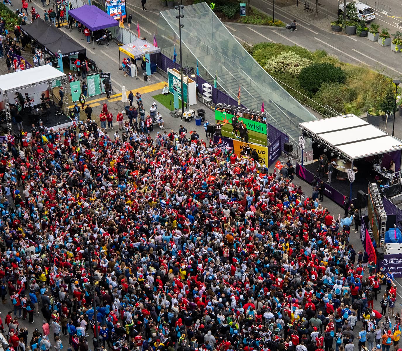 Aerial view of a lively crowd gathered at an outdoor event. People in colorful attire gather near a stage with activities and booths, creating a festive atmosphere.