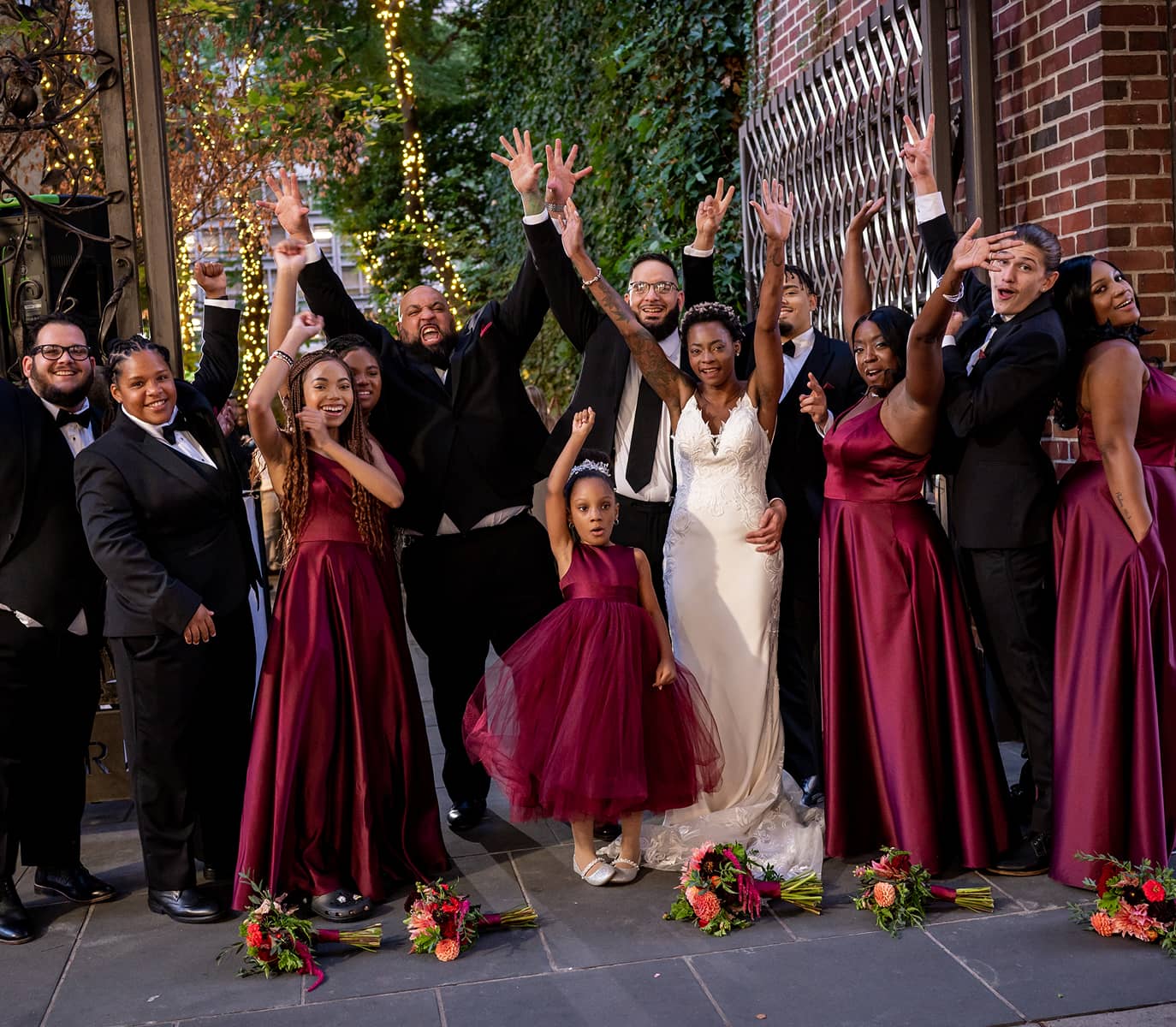 A joyous wedding party poses outside; the bride in a white gown and others in burgundy dresses and suits, arms raised in celebration.