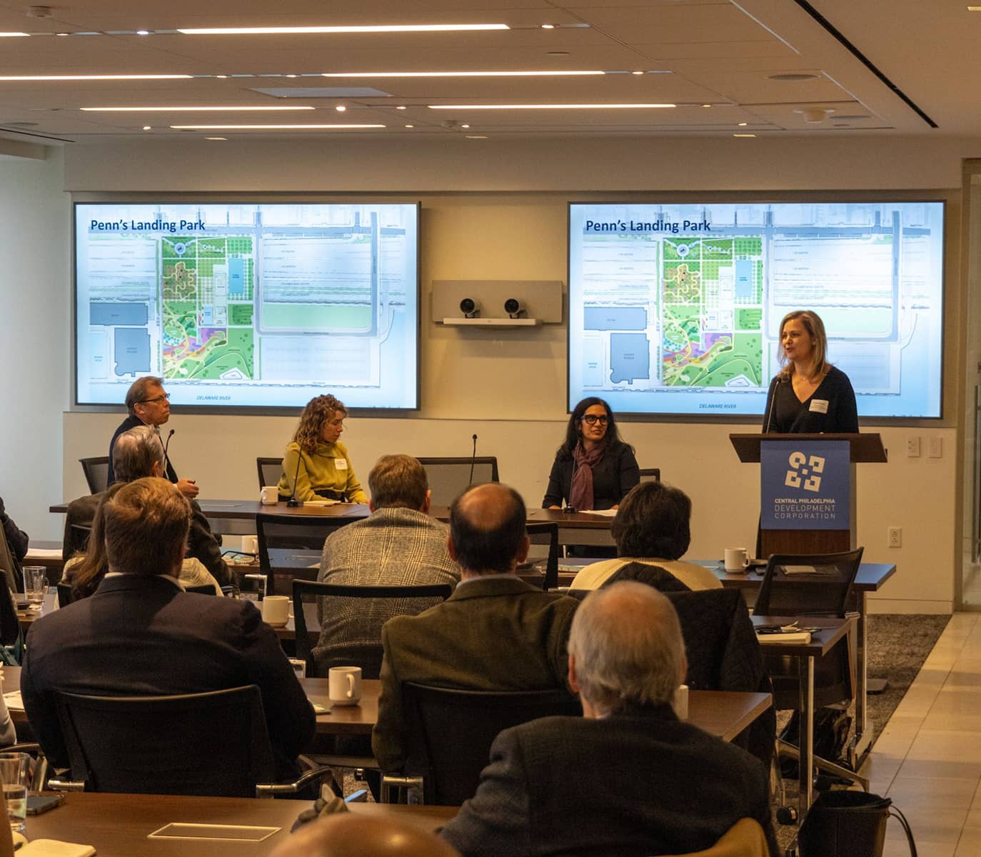 Group presentation in a conference room about Penn's Landing Park. Attendees are seated, two large screens show park plans, and one person speaks at a podium.