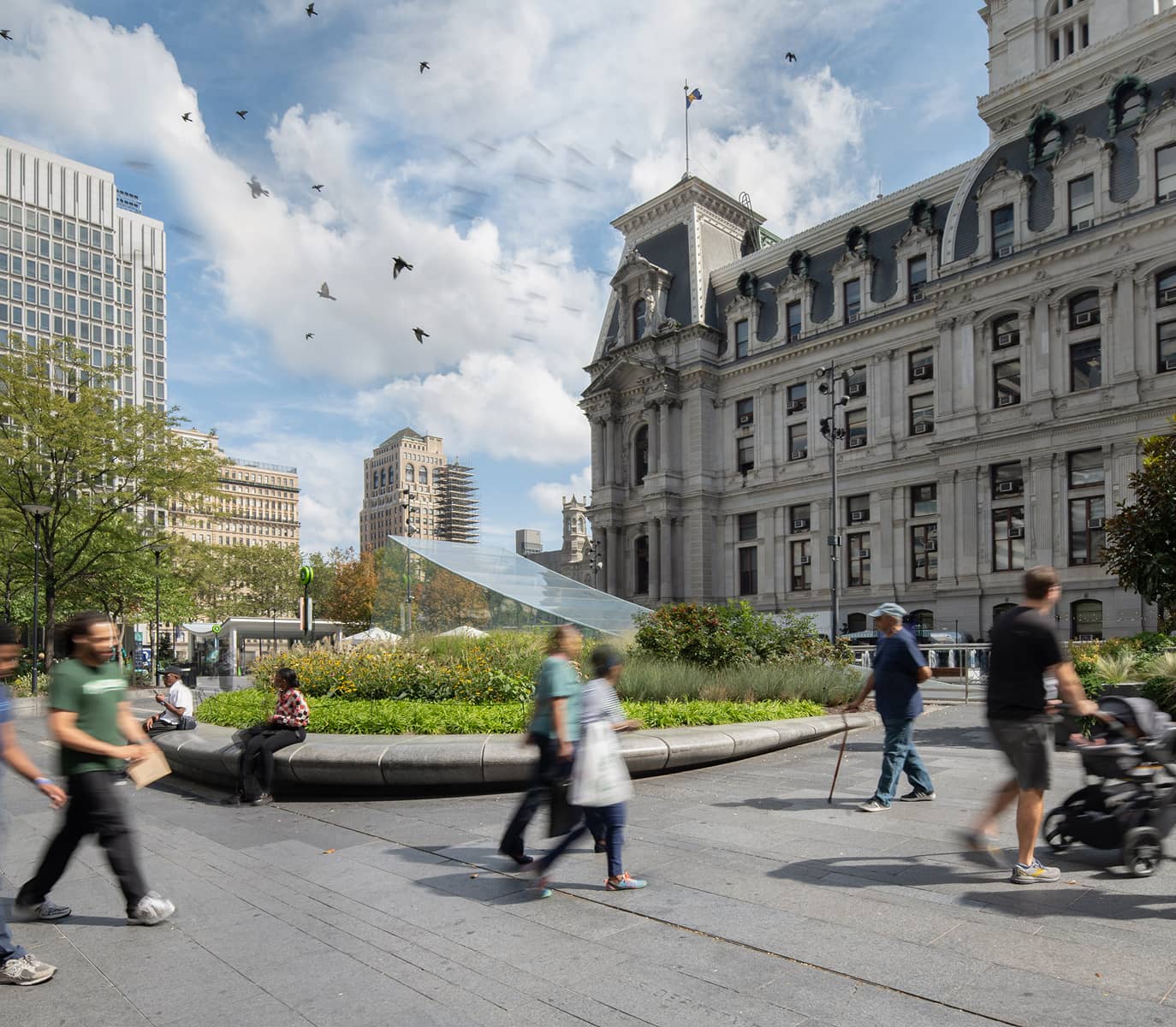 People walking in a bustling urban plaza beside an ornate historic building on a bright day. Birds fly in a blue sky, trees and plants add greenery.