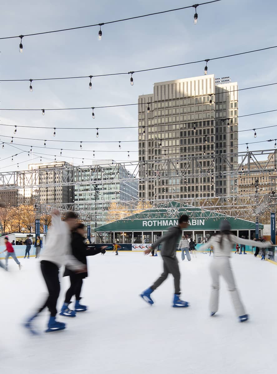 People ice skate on a sunny day at an outdoor rink with string lights above. The 
