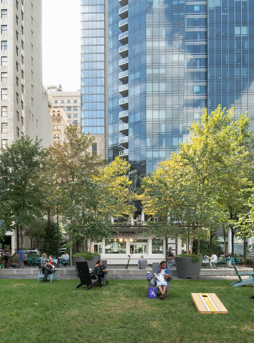 A park scene with people relaxing on chairs and benches, surrounded by green trees. In the background, a modern glass building reflects the sky. The atmosphere feels calm and leisurely.