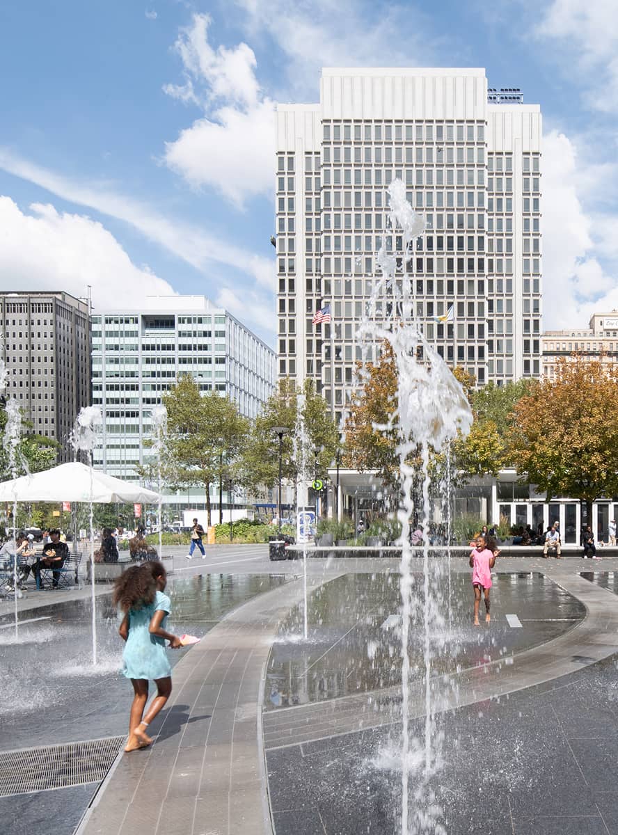 Children play joyfully in a fountain plaza with water jets. Skyscrapers and autumn-colored trees form the background under a bright, blue sky.
