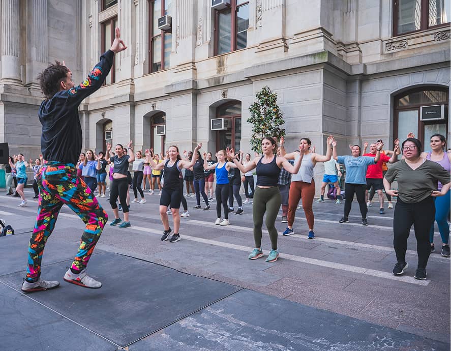 An instructor in colorful leggings leads a lively outdoor workout with a large group of diverse participants. They're exercising in a historic urban setting.