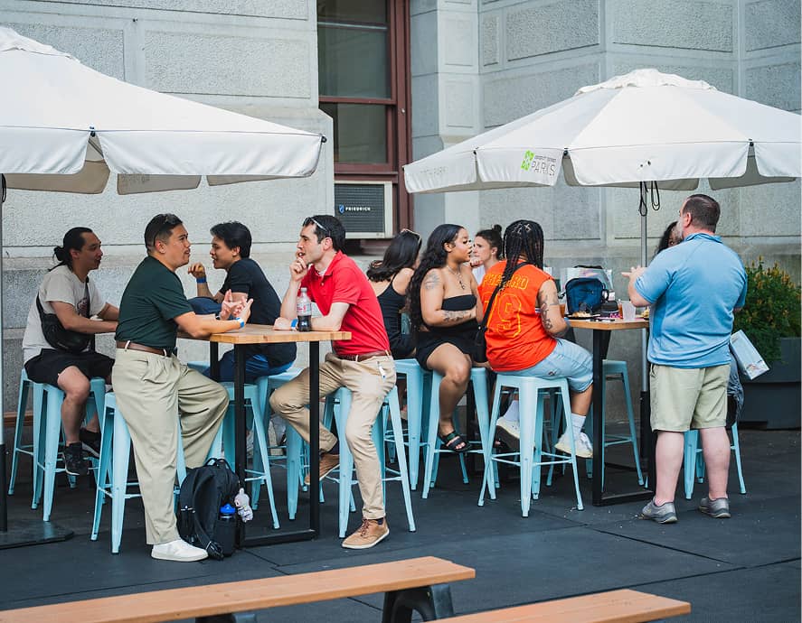People sitting at outdoor tables under umbrellas, engaged in lively conversations. The setting is casual, with a social and relaxed atmosphere.