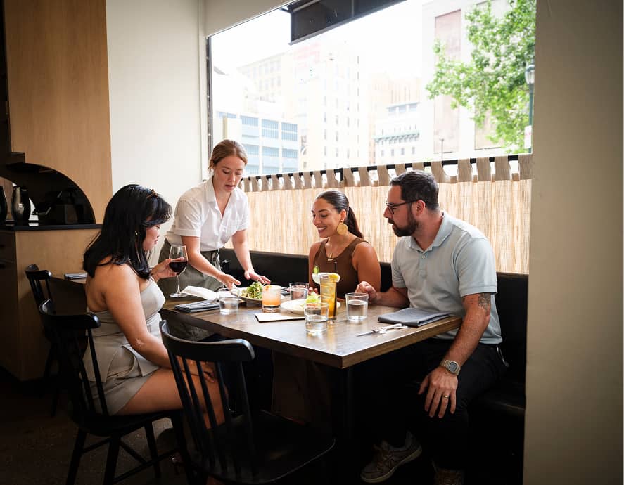 A server places a salad on a table with three smiling diners at a cozy restaurant. A woman holds a glass of red wine, and another has a cocktail. Bright and lively atmosphere.