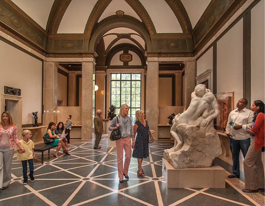 A museum gallery with people observing sculptures. A prominent marble statue shows an embracing couple. The space is grand, with high arches and natural light.