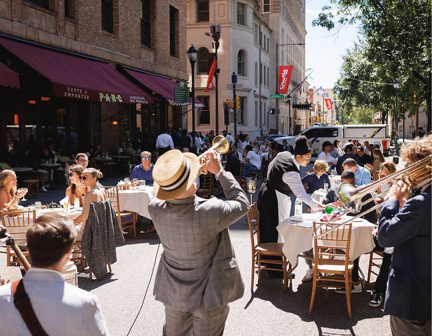 Outdoor street cafe with people dining at tables. A musician in a straw hat plays trumpet, creating a lively, festive atmosphere under sunny skies.