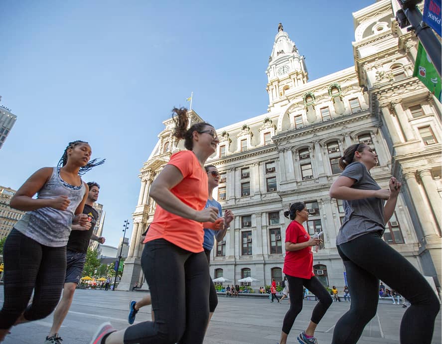 A group of people jog in front of a historic building in an urban setting. They wear colorful athletic gear under a clear blue sky, conveying energy and movement.