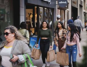 Three women walk with shopping bags in hand, smiling outside an Allbirds store. A bustling city street scene conveys a lively, casual atmosphere.