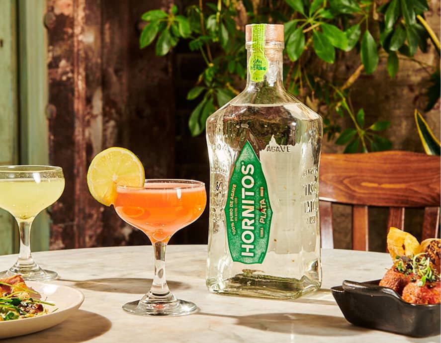 A bottle of Hornitos Plata tequila next to two cocktail glasses, one with an orange drink and lime garnish. The setting is a cozy, warmly-lit dining table with plants in the background.