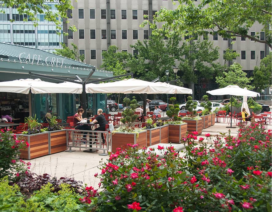 Outdoor café with red flowers and green plants in wooden planters. People sit under white umbrellas, surrounded by urban buildings and trees.