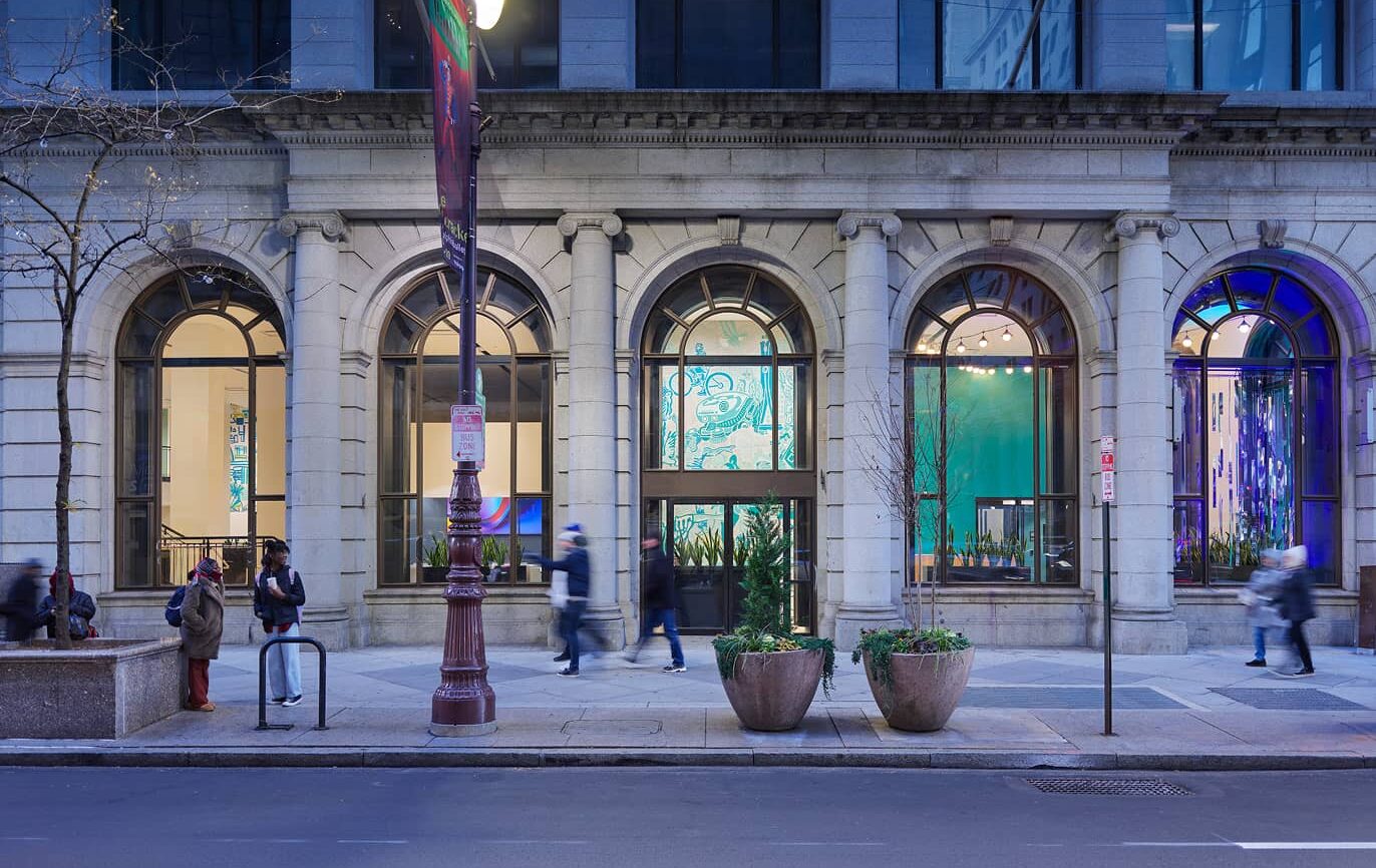 Street view of an illuminated historic building with arched windows. People walk past blurred by motion. The scene is calm with cool blue tones.