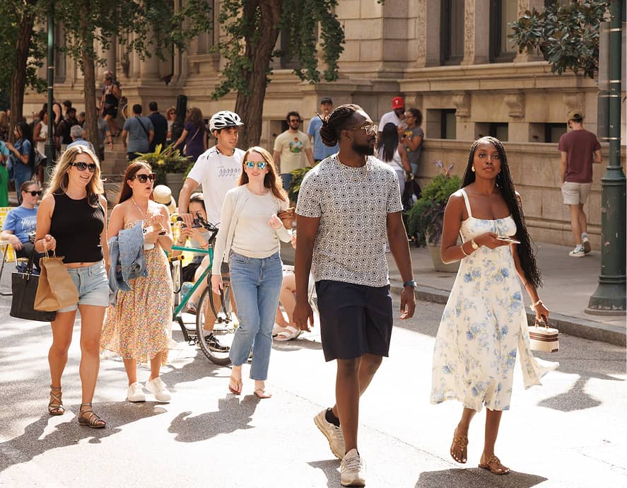 A group of people walk down a sunny city street, smiling and chatting. Trees and a historic building are in the background, creating a casual, lively atmosphere.