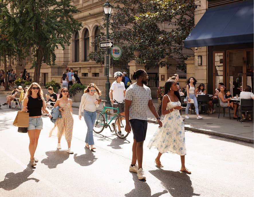 A lively street scene with people walking and a cyclist in an urban setting. Sunlit, casual atmosphere with outdoor seating and lush trees lining the sidewalk.