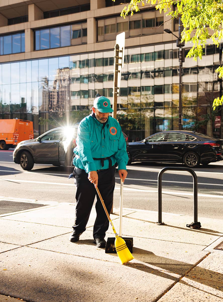 A city worker in a teal uniform sweeps a sidewalk in bright sunlight. Behind him are parked cars, a modern glass building, and a tree with green leaves.