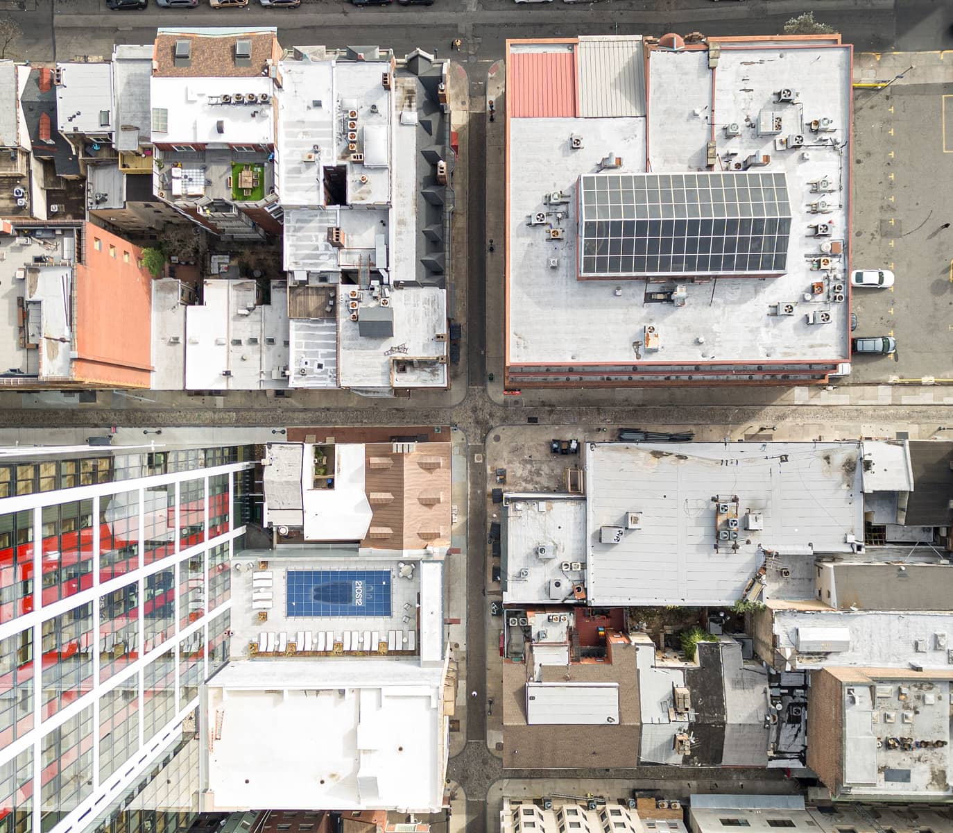 Aerial view of a city block with flat-roofed buildings, solar panels, and a rooftop pool. Streets divide the structures in a grid pattern, creating an urban feel.