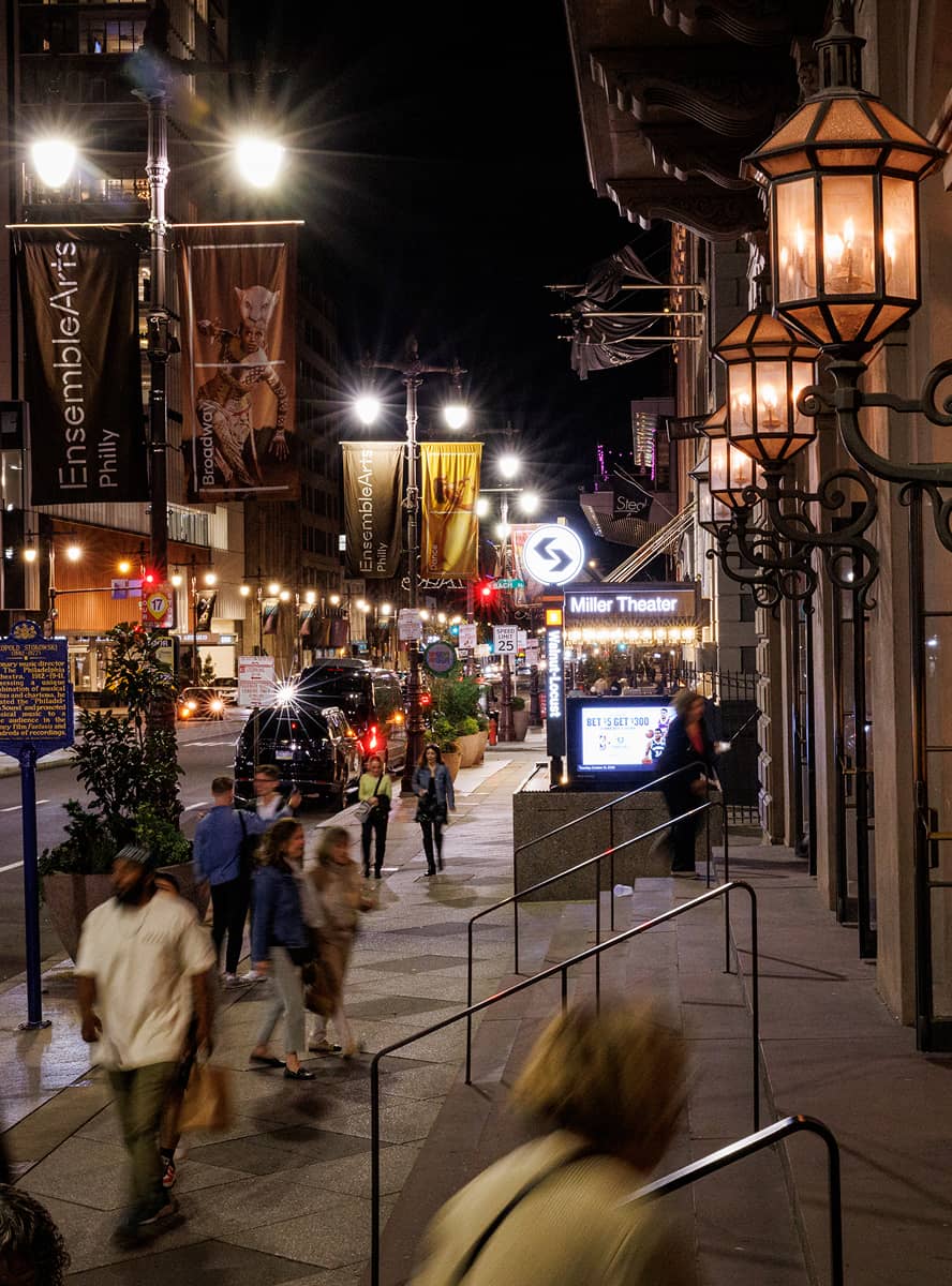 Night scene in a city with people walking along a bustling street. Bright streetlights and banners illuminate the sidewalk near the Miller Theater.