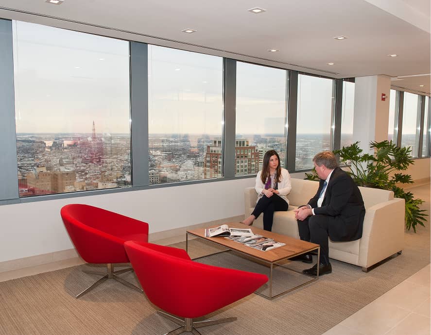 Two people in formal attire sit on a beige couch in a modern office with large windows. Red chairs and plants decorate the space, emitting a professional tone.