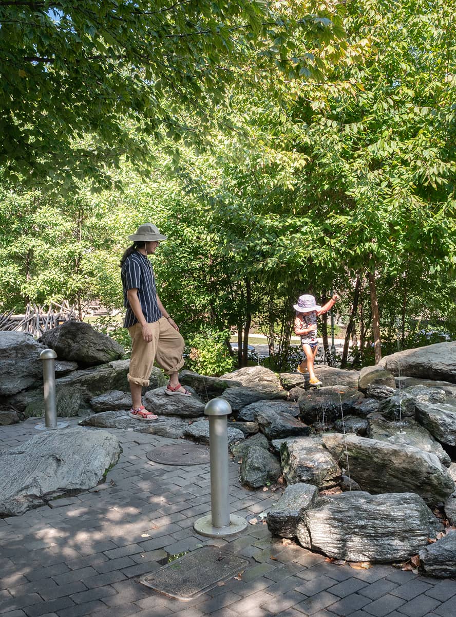A man and a child, both wearing hats, explore a rocky area surrounded by lush green trees. The child balances on rocks, conveying a sense of adventure.