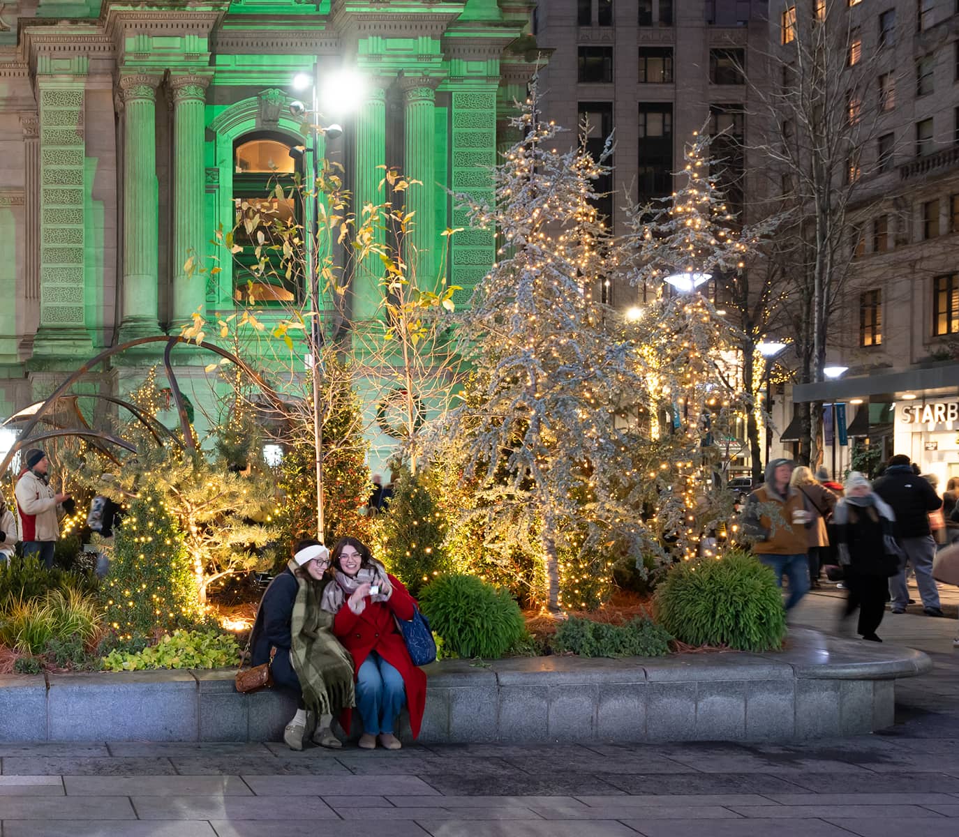 Two people sit on a bench in festive winter attire, surrounded by trees adorned with twinkling lights, in front of a building lit in green.