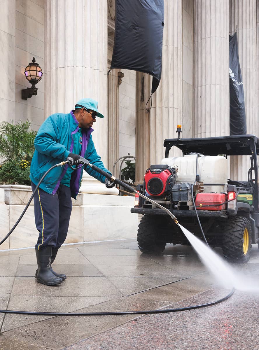A man wearing a turquoise jacket and cap power washes a sidewalk near a building with tall columns. The scene is industrious and focused.