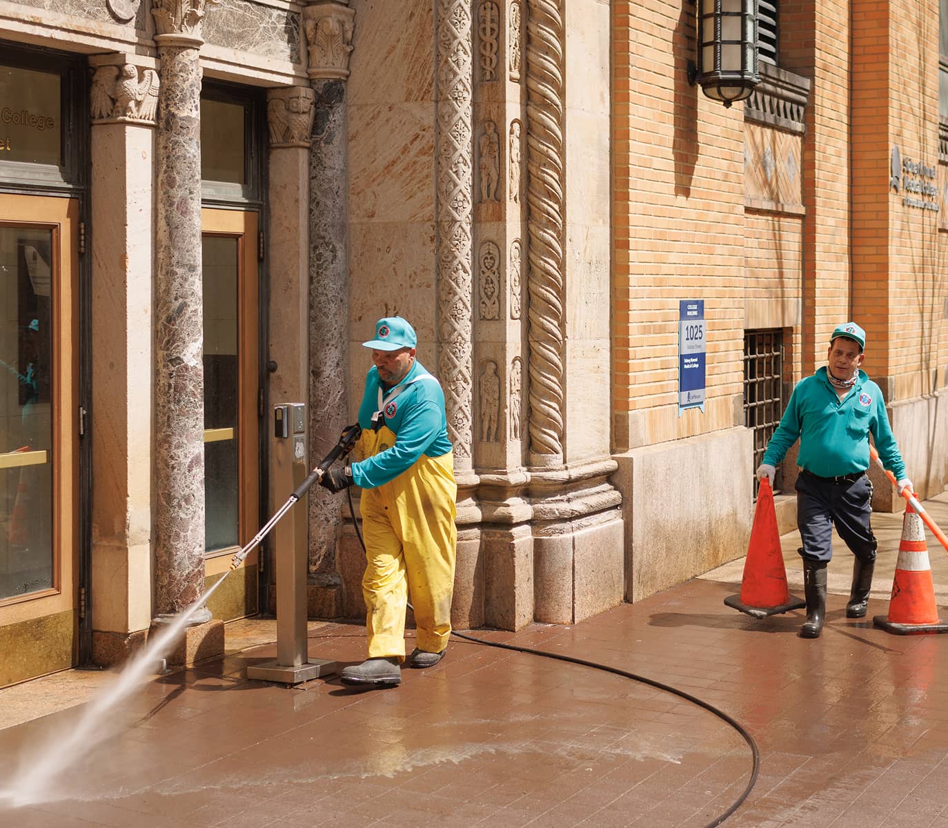 Two workers in teal uniforms and caps clean a sidewalk outside an ornate brick building. One uses a power washer, while the other sets up orange cones.