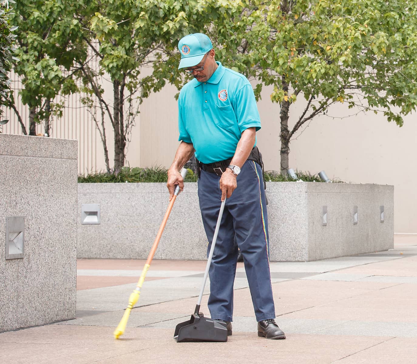 Man sweeping a plaza lined with trees and stone planters. He wears a teal uniform and cap, focused on his task, conveying diligence and calm.