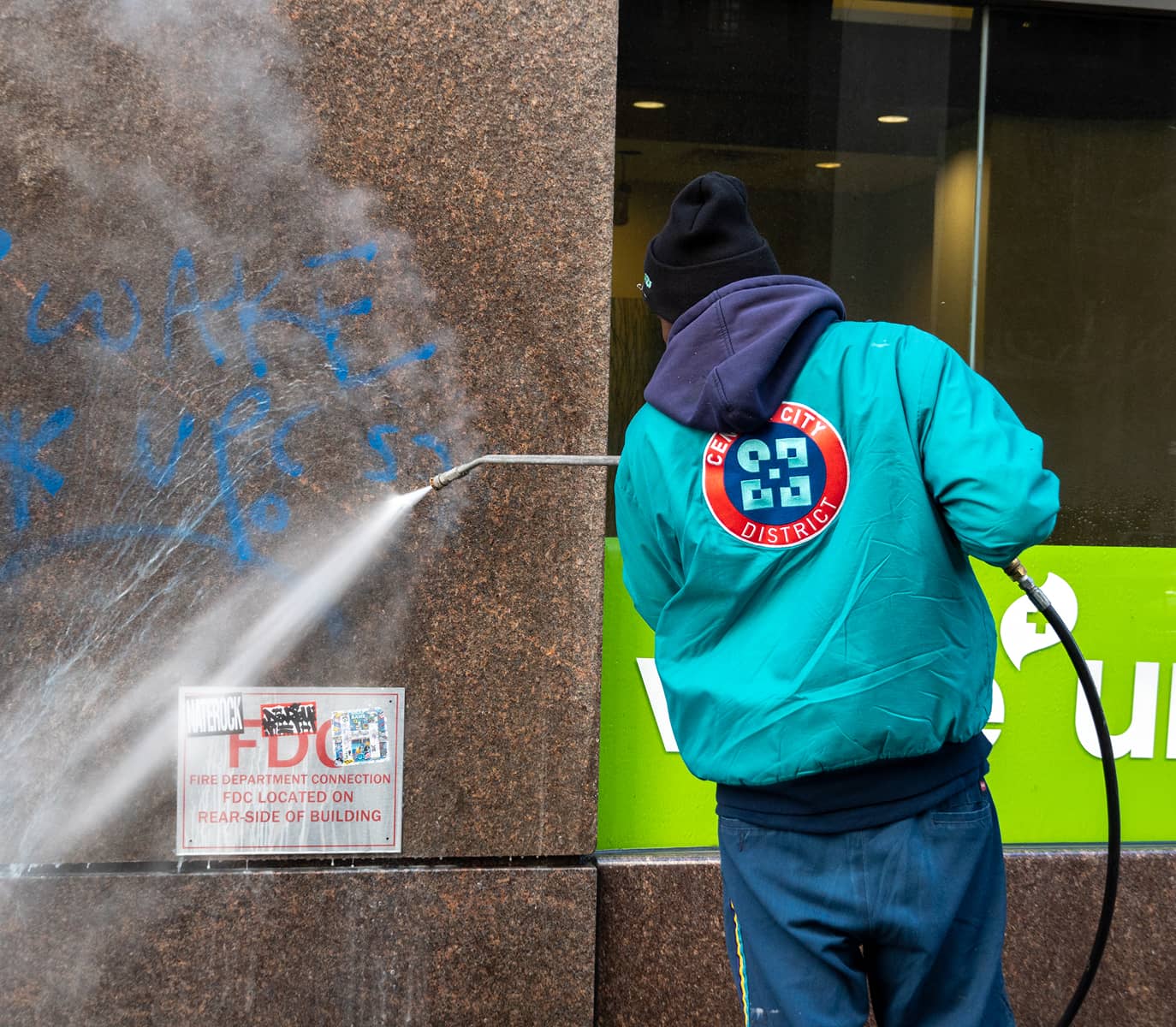 A person in a blue-green jacket power washes blue graffiti off a brown stone wall. The jacket has a red circular logo. The scene conveys urban cleanup.