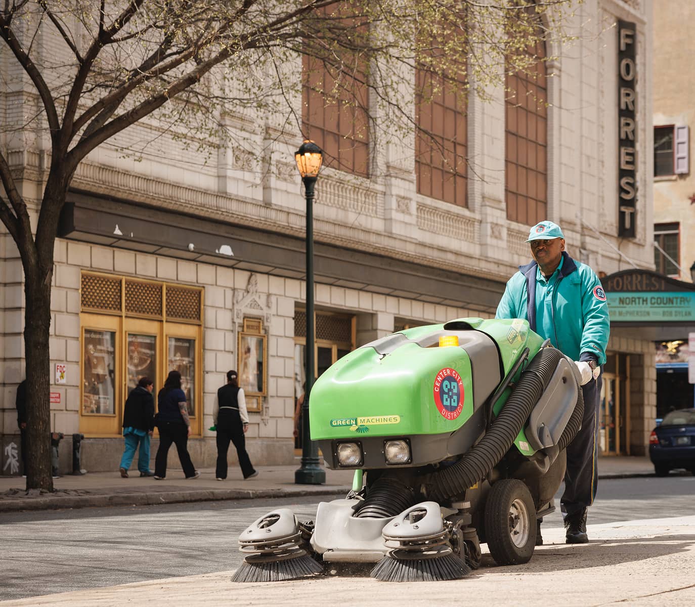 A street cleaner operates a green machine, sweeping a city sidewalk. Nearby buildings and a theater marquee are visible, with pedestrians walking by.