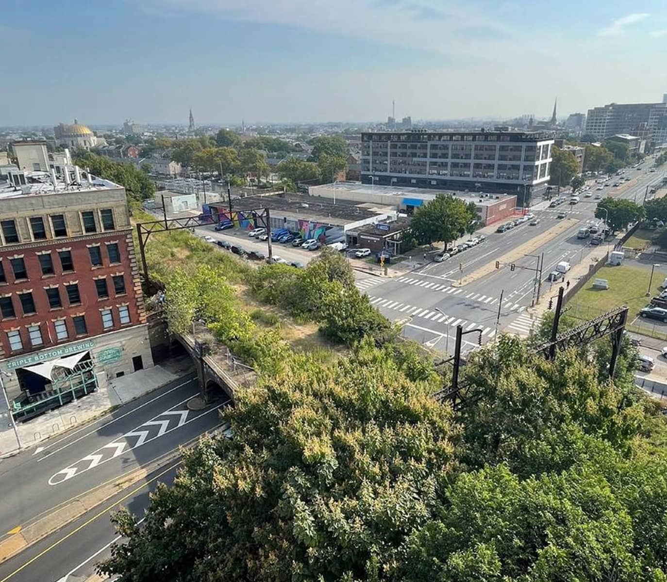 Aerial view of an urban landscape with greenery in the foreground, a historic red-brick building, busy intersection, and modern structures under a clear sky.