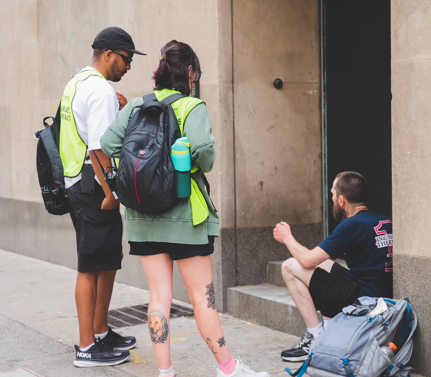 Two people in bright vests with backpacks talk to a seated man at a doorway, seeming to offer assistance. The scene conveys empathy and outreach.