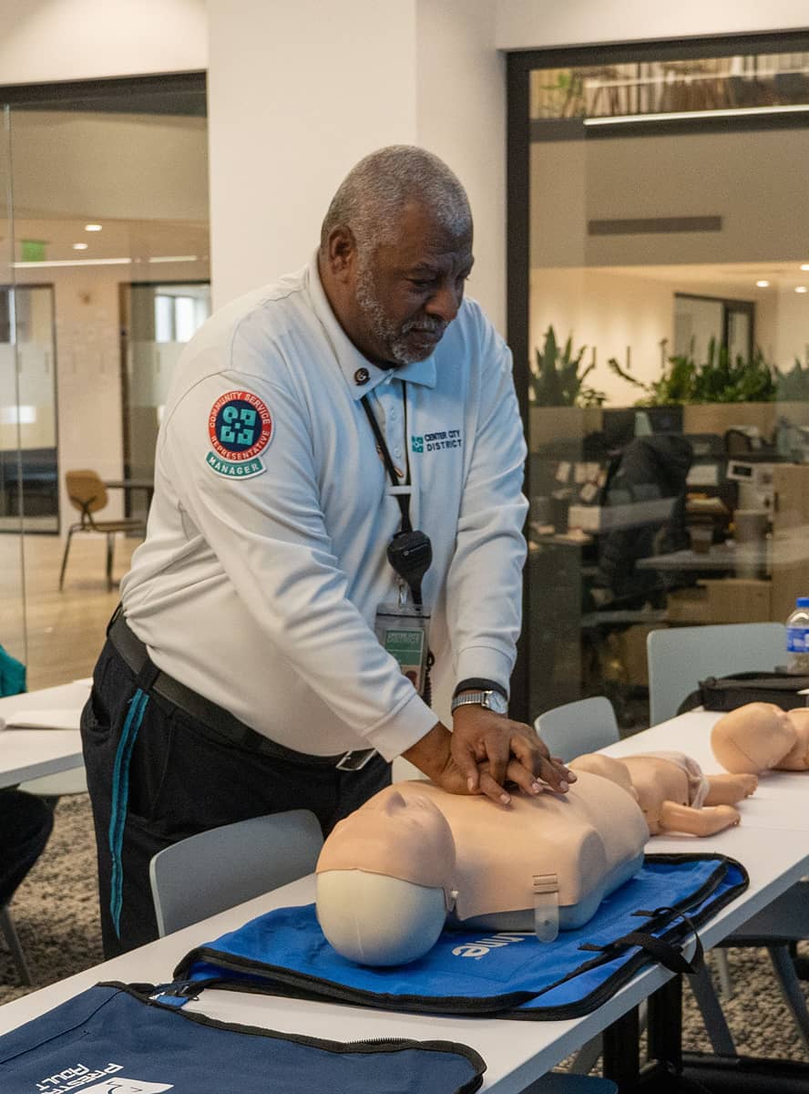 A man demonstrates CPR on a dummy in a classroom setting. He appears focused and professional, with chairs and tables visible in the well-lit room.